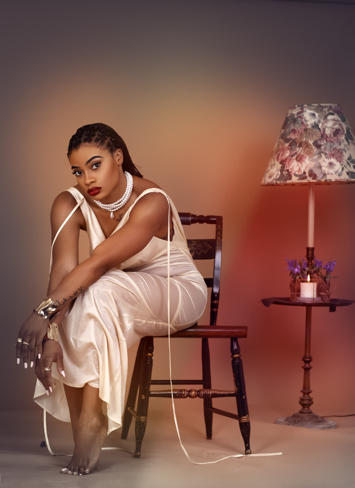 Warm-lit studio portrait of a woman in a cream satin dress and pearls seated beside a vintage lamp